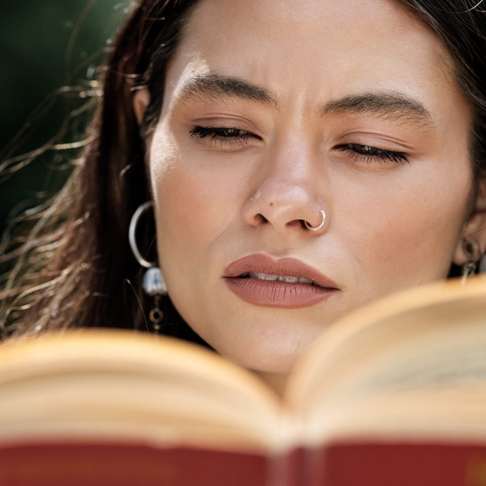 Head-shot of mixed race woman reading a book outdoors. We only can see her relaxed face and the blurry book. Earrings and piercing. Enjoying classic hobbies concept.
